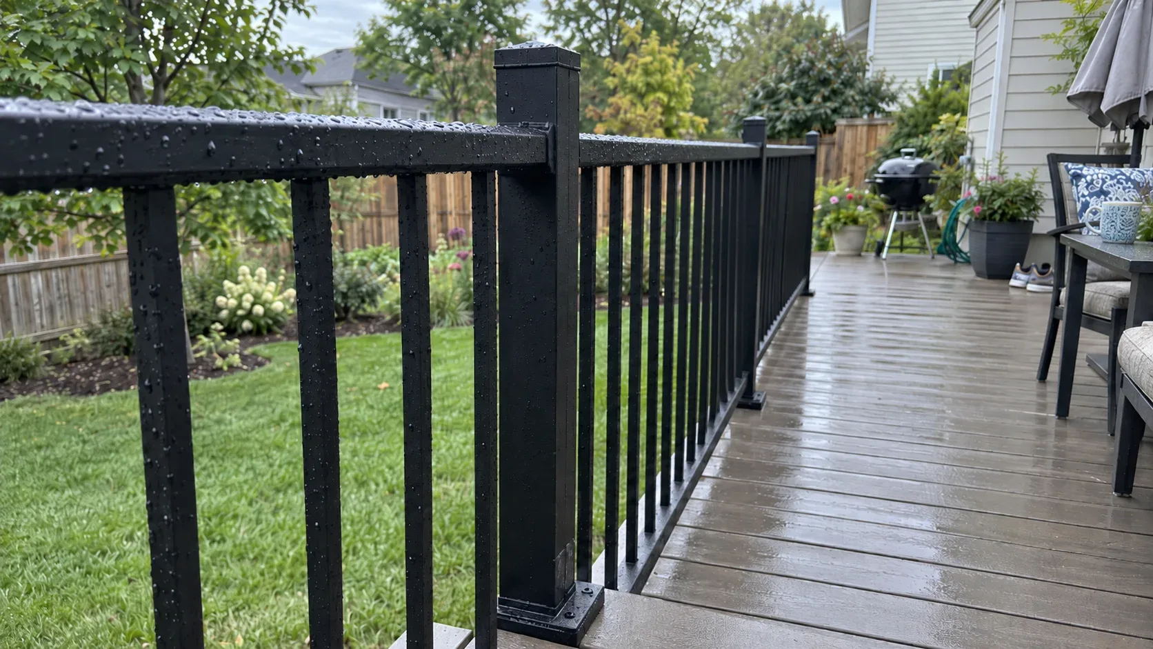 A black aluminium railing beaded with rain, square spindles evenly spaced and the post base plate fixed through the boards